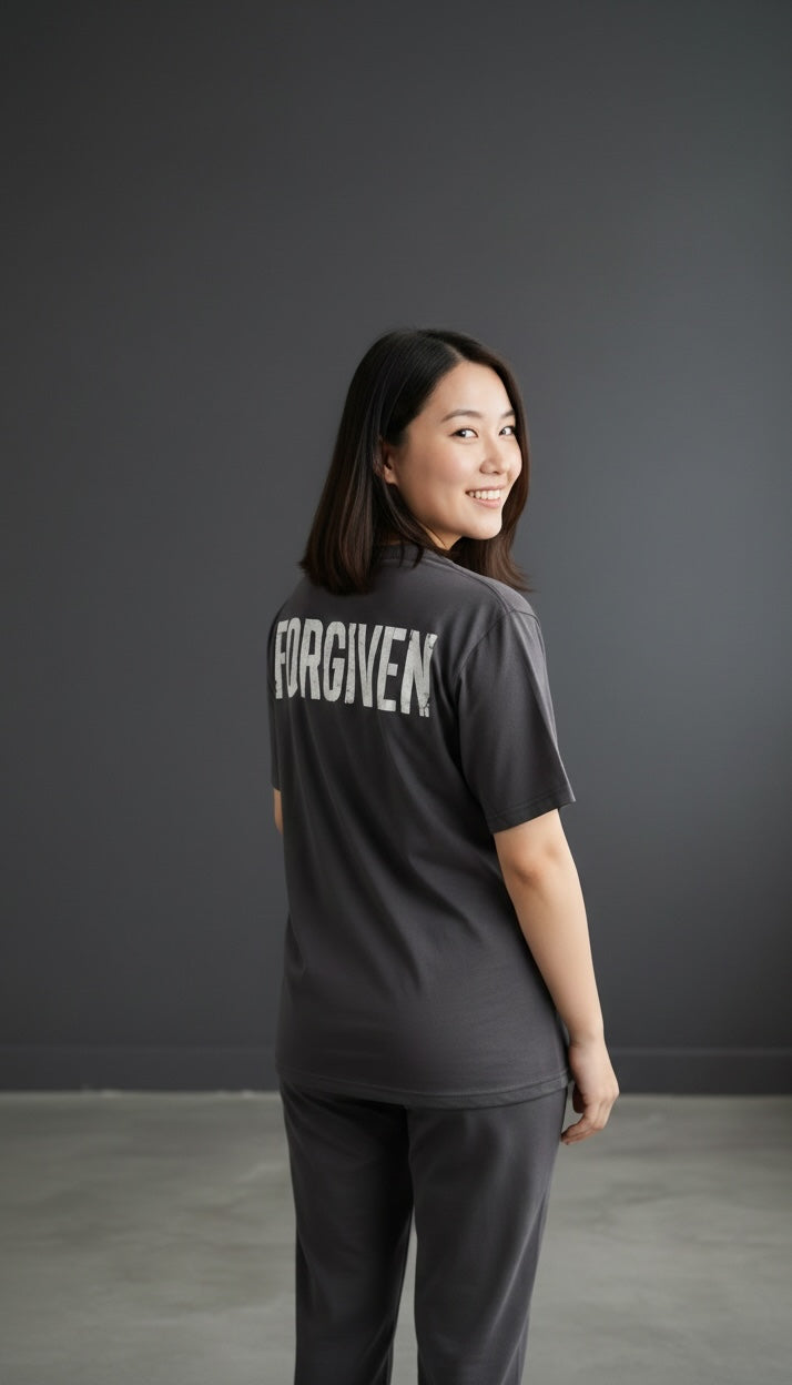 A woman wearing a charcoal grey Forged By God crewneck t-shirt with the word FORGIVEN printed in white on the back.
