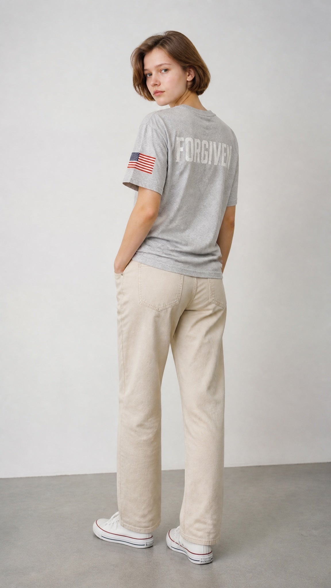 Forged By God grey crewneck t-shirt with FORGIVEN text and US flag sleeve detail worn by a woman with beige jeans