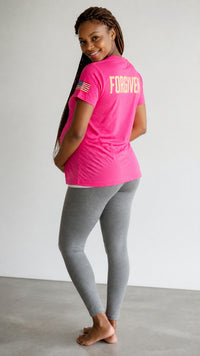 A pregnant woman wearing a bright pink Forged By God crewneck t-shirt with Forgiven printed on the back and grey leggings